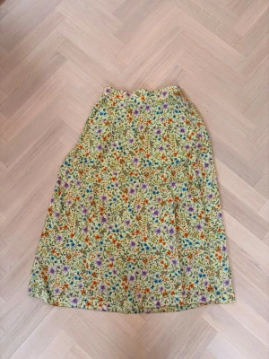 Blommig långkjol i bomull - Söt långkjol med blommigt mönster i flera färger på ljusgrön botten. Kjolen har en avslappnad passform och resår i midjan för extra komfort. Perfekt för dig som gillar färgglada och somriga plagg. 