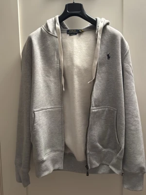 Grå hoodie från Polo Ralph Lauren - Snygg grå zip-hoodie från Polo Ralph Lauren med huva och dragsko. Klassisk logga broderad på bröstet och två stora fickor framtill. Tillverkad i mjukt sweatshirtmaterial som är perfekt för chill dagar. Enkel och stilren design som funkar till det mesta.