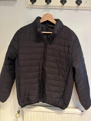 Svart Vår/Höst jacka - Säljer en svart pufferjacka med quiltad design och hög krage. Jackan har dragkedja framtill och två sidofickor. Perfekt för kalla dagar och enkel att matcha med olika outfits. Materialet är syntet och jackan har en klassisk, stilren look.