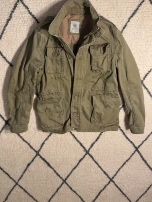 Olivgrön fieldjacket från Marwin - Snygg olivgrön utilityjacka från Marwin med flera stora fickor framtill, axelklaffar och hög krage. Jackan har en avslappnad passform och stängs med knappar. Perfekt för dig som gillar en militärinspirerad look. Ser ut som en Ralph Lauren jacka fast snyggare. 