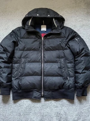 J Lindberg Jacka - Säljer en svart pufferjacka med huva och dragkedja framtill. Jackan har två snedställda fickor med dragkedja och en extra ficka på ärmen. Ribbstickade muddar vid ärmslut och nederkant ger en schysst passform. Perfekt för kalla dagar. I storlek L men passar bättre M. Skriv om ni har funderingar!