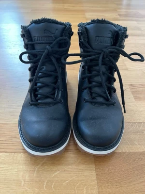 Svarta vinterkängor från Puma - Snygga svarta kängor från Puma med högt skaft och grov sula i vitt. Kängorna har svart snörning och mjukt, fluffigt foder för extra värme. Perfekta för kalla dagar och ger en sportig look till din outfit.