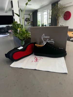 Svarta sneakers från Christian Louboutin - Snygga svarta sneakers från Christian Louboutin med klassisk röd sula och diskret logga. Skorna har lågt skaft, svarta snören och ovandel i mocka. Perfekta för dig som vill ha en exklusiv och stilren look med en ikonisk detalj.