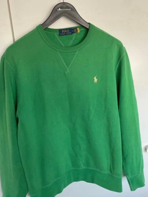 Grön sweatshirt från Polo Ralph Lauren - Snygg grön sweatshirt från Polo Ralph Lauren med klassisk rund halsringning och gul broderad logga på bröstet. Tröjan har ribbade muddar vid ärmslut och nederkant samt diskreta sömdetaljer vid halsen. Perfekt för en avslappnad och stilren look.
