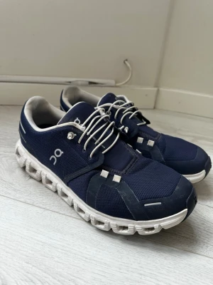 Marinblå On Running sneakers - Snygga marinblå sneakers från On Running med vit sula och elastiska snören. Ovandelen är i meshmaterial för bra ventilation och komfort. Skorna har en sportig design med logga på sidan och detaljer i grått och vitt. Perfekta för dig som gillar stilrena och funktionella sneakers.