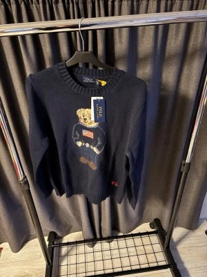 Mörkblå stickad tröja Polo Bear - Mörkblå stickad tröja från Polo Ralph. Tröjan har rund halsringning och ribbade muddar. Perfekt för dig som gillar klassisk och lekfull stil.