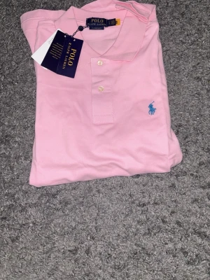 Rosa pikétröja från Polo Ralph Lauren - Snygg rosa pikétröja från Polo Ralph Lauren med klassisk krage, korta ärmar och blå broderad logga på bröstet. Tröjan är i bomull och har knappar framtill. Perfekt för en stilren och avslappnad look.