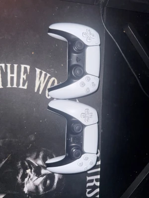 PlayStation 5 DualSense handkontroller - Två PlayStation 5 DualSense handkontroller i vit och svart design. Ergonomisk form med klassiska PlayStation-knappar, touchpad och inbyggd mikrofon. Tillverkade i plast med detaljer i svart och vitt, perfekt för gaming på PS5.