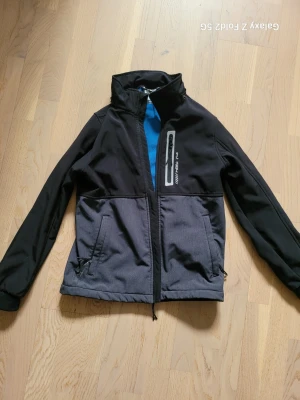 Svart och grå vindjacka från McKinley - Snygg vindjacka från McKinley i svart och grått med blå fleecefoder. Jackan har dragkedja framtill, två sidofickor med dragkedja och en bröstficka med reflexdetalj. Perfekt för blåsiga dagar och outdoor-aktiviteter.