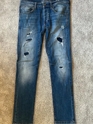slim jeans med slitningar - Säljer ett par blå jeans med flera slitningar och hål på framsidan för en trendig, avslappnad look. Jeansen har klassisk femficksdesign, normal midja och raka ben. Tillverkade i denim med snygg tvätt och slitna detaljer.
