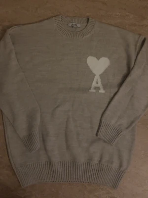 Beige stickad tröja från Ami Paris - Säljer en beige stickad tröja från Ami Paris med ikoniskt vitt hjärta och bokstaven A på bröstet. Tröjan har rund halsringning, ribbade muddar och nederkant. Perfekt för dig som gillar stilrena och trendiga plagg. Storleken är XL men passar dig som har M-L storlek eftersom den har krympit lite.
