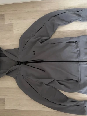 Grå hoodie från BOSS med dragkedja - Snygg grå hoodie från BOSS med hel dragkedja framtill och svarta detaljer. Hoodien har två fickor med dragkedja den är knappt använd helt ny