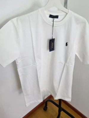 Vit t-shirt från Polo Ralph Lauren - Klassisk vit t-shirt från Polo Ralph Lauren med den ikoniska broderade loggan på bröstet. T-shirten har rund hals, korta ärmar och är gjord i mjuk bomull för en skön känsla. Perfekt basplagg med clean look och snygg passform. Storlek: Large