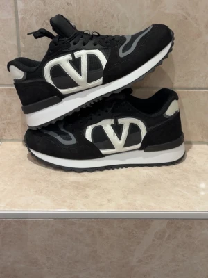 Svarta sneakers från Valentino - Snygga svarta sneakers från Valentino med stora vita V-loggor på sidorna. Skorna har en mix av mocka och textil, vita detaljer och svart sula med grå inslag. Klassisk låg modell med snörning och bekväm passform.