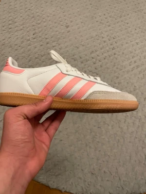 Addidas samba sneaker   - Säljer ett par vita Adidas sneakers med klassiska rosa ränder på sidan och beige mockadetalj vid tån. Skorna har en låg siluett och gummisula i brunt. Perfekta för dig som gillar retrostil och vill ha något som sticker ut lite extra.