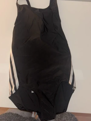 Svart baddräkt från Adidas - Klassisk svart baddräkt från Adidas med vita ränder längs sidorna.  