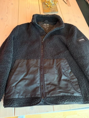 Svart teddyjacka från Calvin Klein - Mysig svart teddyjacka från Calvin Klein med dragkedja framtill. Jackan har stora fickor i nylonmaterial och är gjord av återvunnen teddy fleece och nylon. Hög krage och diskret logga på ärmen ger en clean och modern look. Storlek M