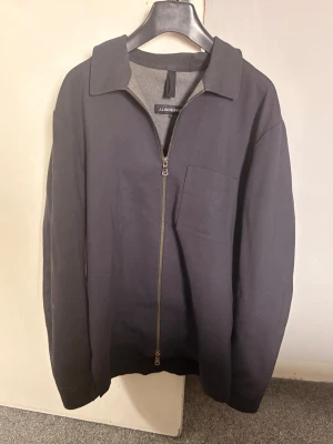 Mörkblå overshirt från J.Lindeberg - Stilren mörkblå overshirt från J.Lindeberg med rak passform, klassisk krage och bröstficka. Dragkedja framtill i metall och mjukt bomullsmaterial. Perfekt lager på lager-plagg för en clean look. i armhålan har den  öppnats upp på insidan men ingenting som syns utanpå.