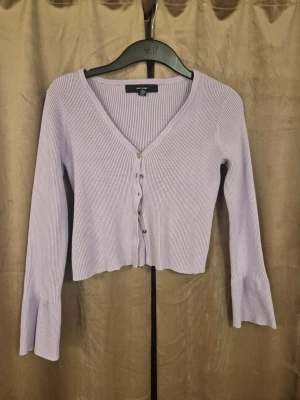 Ljuslila ribbad kofta från H&M - Trendig ljuslila kofta från H&M med ribbad struktur, v-ringning och knappar framtill. Modellen är croppad med långa ärmar och lätt utsvängda ärmslut. Perfekt att styla över ett linne eller t-shirt för en chill look.