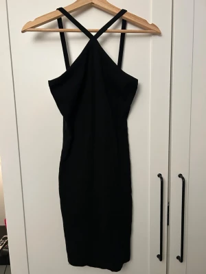 Svart tight klänning med korsade band - Svart klänning med kors i fram. Modellen är tight och har en enkel, stilren design.
