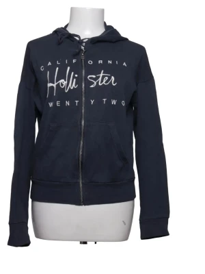 Marinblå hoodie från Hollister - Snygg marinblå hoodie från Hollister med dragkedja framtill och vit text på bröstet. Hoodien har huva med dragsko och två fickor fram. Tillverkad i mjukt bomullsmaterial som känns skönt mot huden. Perfekt för en avslappnad och cool stil.