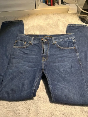 Mörkblå jeans från Nudie Jeans - Säljer ett par klassiska mörkblå jeans från Nudie Jeans med raka ben och fem fickor. Jeansen har kontrastsömmar i gult och är tillverkade i slitstark denim. Perfekta för dig som gillar en tidlös och enkel stil. 8,5/10