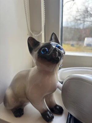 Porslinskatt med blå ögon - Söt liten porslinskatt i beige och svart med stora blå ögon. Katten har en blank yta och är formad i en sittande position. Perfekt som dekoration i fönstret eller på hyllan.