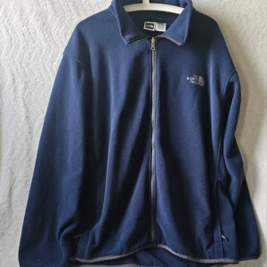Marinblå fleecejacka The North Face - Mysig marinblå fleecejacka från The North Face med dragkedja framtill och hög krage. Jackan har broderad logga på bröstet och två sidofickor med dragkedja. Perfekt för lager-på-lager och chill dagar.