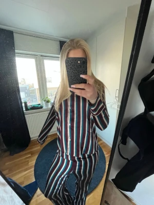 Randig pyjamas i satinlook - Snygg tvådelad pyjamas med långa ärmar och byxor i satinliknande material. Setet har vertikala ränder i vinrött, svart, vitt och turkost. Skjortan har knappar framtill och klassisk krage. Perfekt för mysiga kvällar hemma och skönt att sova i under den kalla perioden. 