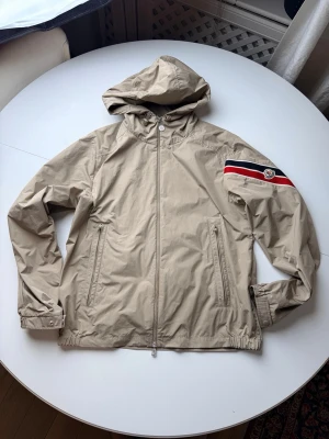 Beige Moncler vindjacka med huva - Snygg beige vindjacka från Moncler med huva och dragkedja framtill. Jackan har två fickor med dragkedja och en cool detalj på ärmen med Moncler-logga och röd, vit och blå rand. Perfekt för att hålla vinden borta och se stylish ut.