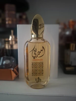 Ameerati parfym från Al Wataniah - Ameerati parfym från Al Wataniah i en elegant, genomskinlig glasflaska med guldfärgat lock och detaljer. Flaskan har arabiska inskriptioner och ett dekorativt mönster på framsidan. Doften är exklusiv och flaskan ger en lyxig känsla.