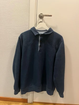 Mörkblå collagetröja med krage - Mörkblå collagetröja från Allan Clark med klassisk krage och knappslå framtill. Tröjan har långa ärmar, ribbade muddar och en avslappnad passform. Perfekt för en chill och stilren look.