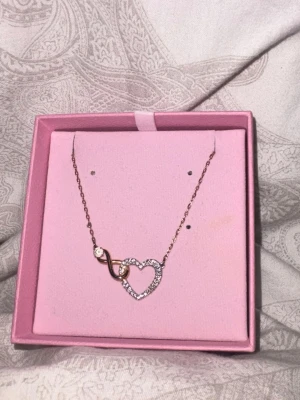 Swarovski halsband med hjärta - Säljer ett elegant halsband från Swarovski i roséguld med ett gnistrande hjärthänge dekorerat med små klara stenar och en infinity-symbol. Kedjan är tunn och smycket kommer i original rosa ask med svanlogga. Perfekt för dig som gillar stilrena detaljer. Pris kan diskuteras.