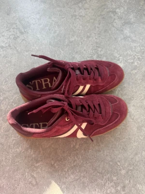 Vinröda sneakers från Stradivarius - Snygga vinröda sneakers från Stradivarius med klassiska vita ränder på sidan och bruna gummisulor. Skorna har en låg profil, snörning och är tillverkade i mockaliknande material. Perfekta för dig som vill ha en stilren och trendig look.