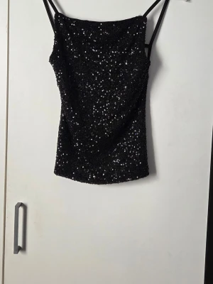 Top från Gina Tricot  - Svart paljettopp med öppen rygg. Paljettoppen är svart och har normal passform. Den har smala axelband och reglerbara resårband i ryggen. 