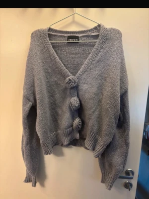 kofta från Pieces - Superfin grå stickad kofta från Pieces med stora dekorativa virkade blommor framtill vid knappar. Koftan har v-ringning, ribbade muddar och en lite croppad passform. Perfekt att slänga över en topp för en mysig look eller till en fin sommarklänning för kyligare dagar. Knappt använd