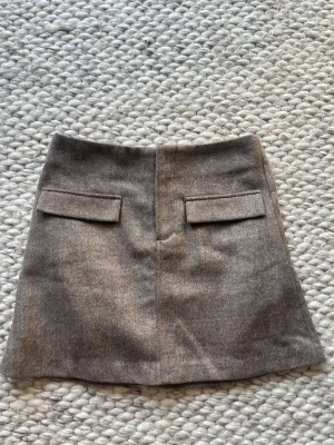 Tailored Skirt Gina tricot  - Snygg beige kortkjol med dekorativa ficklock framtill. Kjolen har en A-linjeformad siluett och neutral färg som är lätt att matcha. Perfekt för dig som gillar stilrena och trendiga plagg.