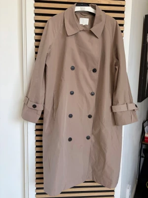 Beige trenchcoat med knappar - Stilren beige trenchcoat med dubbelknäppning och klassisk krage. Jackan har svarta knappar, sidofickor och justerbara ärmremmar. Materialet ser ut att vara bomull eller en bomullsblandning. Perfekt för dig som gillar tidlös och enkel stil.
