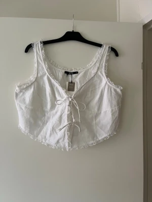 Vit linnetopp med snörning och spets - Söt vit linnetopp med snörning framtill och spetsdetaljer längs kanterna. Toppen är ärmlös och har en v-ringad form som ger en feminin look. Perfekt för varma dagar och enkel att styla med jeans eller kjol.