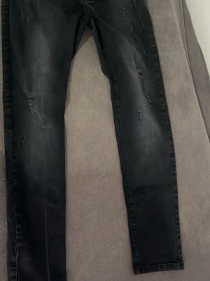 Svarta skinny jeans med slitningar - Säljer ett par svarta skinny jeans med diskreta slitningar framtill. Jeansen har klassisk femficksdesign och smal passform hela vägen ner. Materialet är stretchigt denim som sitter tajt och bekvämt. Perfekta för en trendig och avslappnad look.