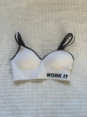 Vit sport-bh med svart text - Sport-bh i vitt med svarta detaljer och texten 'WORK IT' framtill. Justerbara axelband och vadderade kupor för extra stöd. Perfekt för träning och aktiva dagar, den är lite nopprig (se sista bilden)!💓🌸