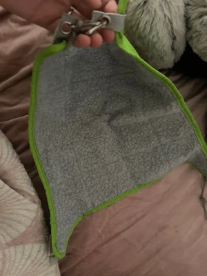 Grå och grön hängmatta till smådjur - Säljer en mjuk hängmatta för smådjur, med grå insida i fleeceliknande material och grön kant. Hängmattan har metallkrokar och öglor för enkel upphängning i bur. Perfekt för exempelvis råttor eller illrar.