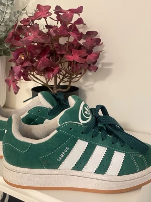 Adidas Campus gröna sneakers - Gröna Adidas Campus sneakers i mocka med klassiska vita ränder på sidorna och vit logga på plösen. Skorna har mörkgröna skosnören, vit mellansula och gummifärgad yttersula. Snygg retrostil som passar till många outfits.