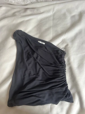 Mörkgrå oneshoulder topp från Zara - Använd en gång. Perfekt inför sommaren snygg mörkgrå oneshoulder topp från Zara med rynkade detaljer på sidan. Toppen är axelbandslös på ena sidan och har en tight passform som framhäver siluetten. 