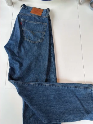 Levi's blå raka jeans - Klassiska blå jeans från Levi's med rak passform och hög midja. Jeansen har fem fickor, Levi's ikoniska läderpatch bak och röd logotagg på bakfickan. Tillverkade i slitstark denim, perfekta för en avslappnad och tidlös stil.