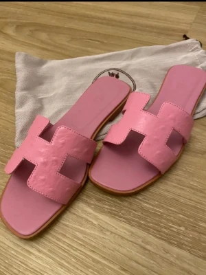 Rosa sandaler med H-design - Säljer ett par trendiga rosa sandaler med öppen tå och breda remmar i form av ett H. Sandalerna har platt sula och är tillverkade i slätt läder. Perfekta för en avslappnad och stilren look.