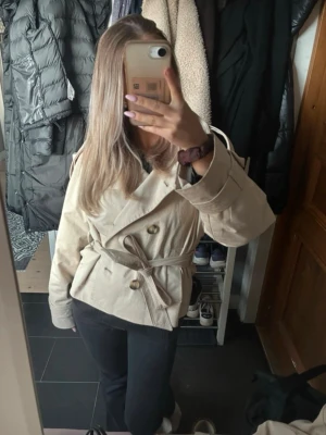 Beige trendig trenchcoat  - Kort beige trenchcoat med dubbelknäppning och bälte i midjan. Jackan har bred krage och dekorativa axelklaffar. Passar perfekt till höst och vår och ger en klassisk look. Materialet ser ut att vara bomull eller en bomullsblandning.