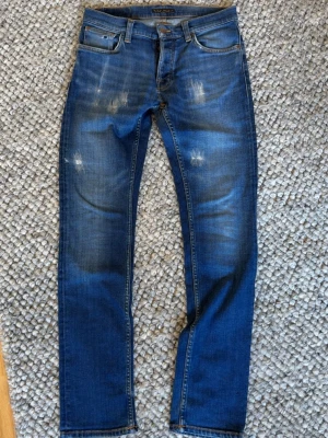 Blå slitna jeans från Nudie Jeans - Snygga blå jeans från Nudie Jeans med slitningar och tvättade detaljer för en cool look. Klassisk femficksmodell med raka ben och normal midja. Tillverkade i denim med kontrastsömmar och läderpatch bak i midjan.