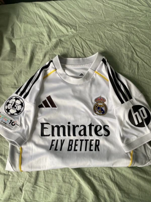 Äkta/original Real Madrid Mbappé matchtröja - Säljer en vit Real Madrid matchtröja från Adidas med Mbappé och nummer 10 på ryggen. Tröjan har svarta detaljer, klubbmärke, Champions League-märke och sponsorloggor som Emirates och HP. Tillverkad i funktionsmaterial, perfekt för fotboll eller som supporterplagg.