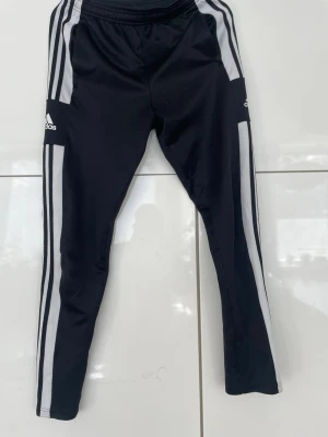 Svarta Adidas mjukisbyxor med vita ränder - Snygga svarta mjukisbyxor från Adidas med klassiska vita ränder längs benen. Tillverkade i Primegreen-material och har en elastisk midja för skön passform. Perfekta för träning eller chill. Adidas-logga på låret och smal passform.